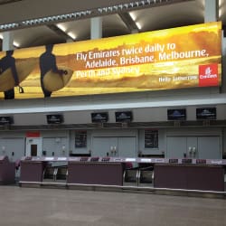 An empty airport check-in counter is bathed in LED lighting. Above the counter, both backlit advertising and illuminated signage for Emirates gleam, promoting twice daily flights to Adelaide, Brisbane, Melbourne, Perth, and Sydney. The Emirates logo stands out brightly in the advertisement.