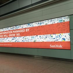 Large SanDisk advertisement on an LED advertising board reads, "Total Information Managed by Enterprises Will Grow 14x." The background is filled with small icons and illustrations related to data and technology. The ad is placed in what appears to be an airport terminal.