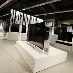 A large, curved flat-screen TV is displayed on a white stand in an electronics store. The background shows several other televisions on display with modern store fixtures and bright overhead lighting, with a bright green beam used to illuminate new retail features for John Lewis.