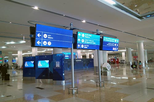 An airport terminal with informational signs hanging from the ceiling. These signs provide directions to various locations, such as the metro, check-in counters, and departure gates, in multiple languages on an LED lit wayfinder.