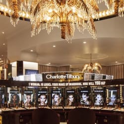 A glamorous, well-lit makeup display in Westfield London showcases various Charlotte Tilbury products. The setup includes large promotional images of models under a sign reading, "Charlotte Tilbury." LED light sheet displays and a luxurious chandelier overhead emphasize the upscale ambiance.