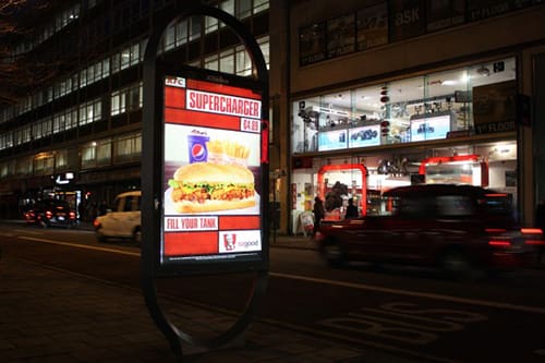 A nighttime street view showcasing a well-lit advertisement display promoting KFC's Supercharger meal, including chicken, fries, and a Pepsi.