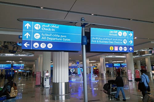 An airport terminal with signs directing passengers to various locations. The blue wayfinders display icons and text in both Arabic and English, indicating directions for "Check-In," "All Departure Gates," "Arrivals," and "First & Business Class Check-In."