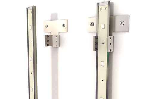 Image of two metallic vertical bars with multiple rectangular slots, mounted on a white wall using brackets. Each bar has screws securing the brackets and slots, likely for adjustable mounting purposes. The setup appears to be part of a modular shelving system or hardware fixture.