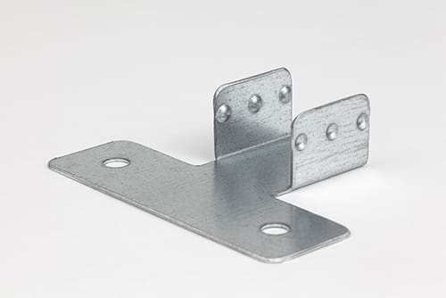 A small metal mounting bracket rests on a white surface. The bracket features a flat base with two large holes and a perpendicular side component with two vertical tabs. Each tab has three smaller holes, intended for attachment or connection purposes.