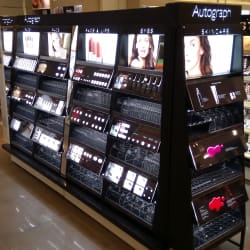 A well-lit LED illuminated makeup stand in a store, showcasing various beauty products including lipsticks, foundations, and eyeshadows. The shelves are organized with product samples and promotional images of models. The display is branded with the name "Autograph.