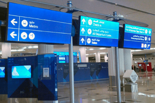 Airport terminal signs in both English and Arabic, illuminated by Dubai airport LEDs, display directions to Metro, Check-in, All Departure Gates, Unaccompanied Minors, and Toilets. The signs hang from the ceiling in the well-lit terminal area with a modern design.