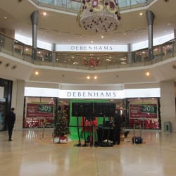 A spacious mall interior with a Debenhams store prominently displayed. The store is decorated with holiday-themed posters advertising a 30% discount. LED retail illumination at Debenhams highlights the festive decor. A Christmas tree and a stage setup with audio-visual equipment are visible in front of the store.