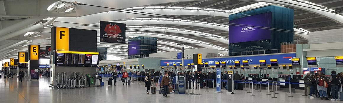 A spacious airport terminal with a high, arching roof. Multiple check-in counters with people in line are visible on the right. Overhead LED wayfinding signage for gates D, E, and F is suspended from the ceiling. A large flight information display board is on the left.