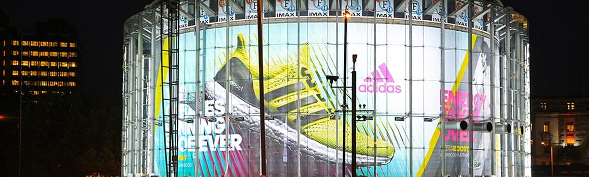 A large illuminated LED matrix on a round building displays a prominent out-of-home advertisement for Adidas shoes. The ad features a vibrant yellow running shoe with black stripes, accompanied by text and the Adidas logo in pink. The background includes various colorful diagonal lines.