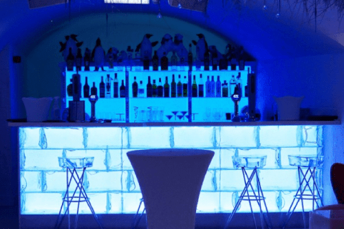 A modern bar featuring blue LEDs and a curved ceiling boasts a counter with a backlit, ice-like design. Bar stools line the counter, and a tall, round cocktail table stands in the center. Shelves behind the bar display assorted bottles, creating an alluring ICE bar ambiance.