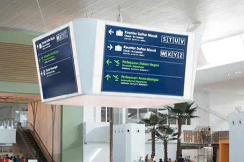 The interior of Kuala Lumpur Airport features LED-illuminated directory signs pointing to services including check-in counters, domestic, and international departures. The signs are in multiple languages. Palm plants decorate the space, and travelers walk in the background.