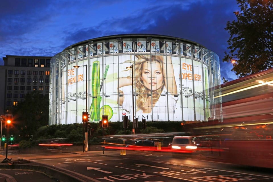 The IMAX, LED illuminated at night, features a giant advertisement for a makeup brand with a woman's face and green mascara. Traffic lights and blurred light trails from moving vehicles can be seen in front of the building. Trees and buildings surround the area.