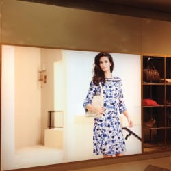 A large fashion advertisement features a woman in a stylish blue and white floral dress standing against a minimalist background. The image is illuminated and displayed on a wall next to shelves with various fashion accessories.