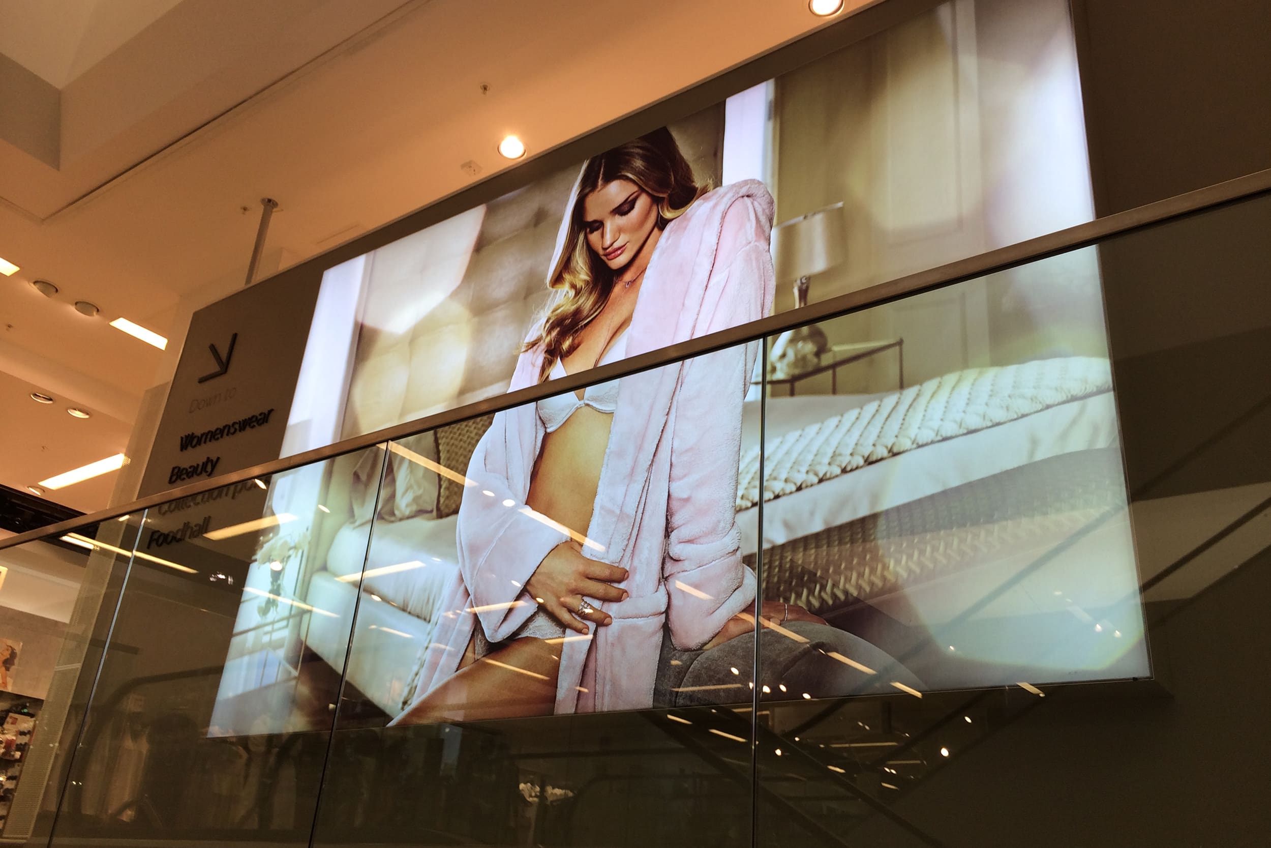 A large LED advertisement in a shopping mall displays a woman wearing a light pink bathrobe over white lingerie, posing in a bedroom setting. The M&S ad is mounted behind a glass railing with store sections such as "Womenswear" and "Beauty" indicated on the left.