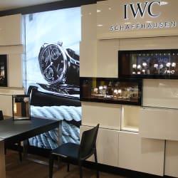 A display at an IWC Schaffhausen store showcasing luxury watches. The setup includes a large screen featuring a close-up image of a watch, glass display cases with watches inside, a black table, and a black chair. The store's logo is prominently displayed.