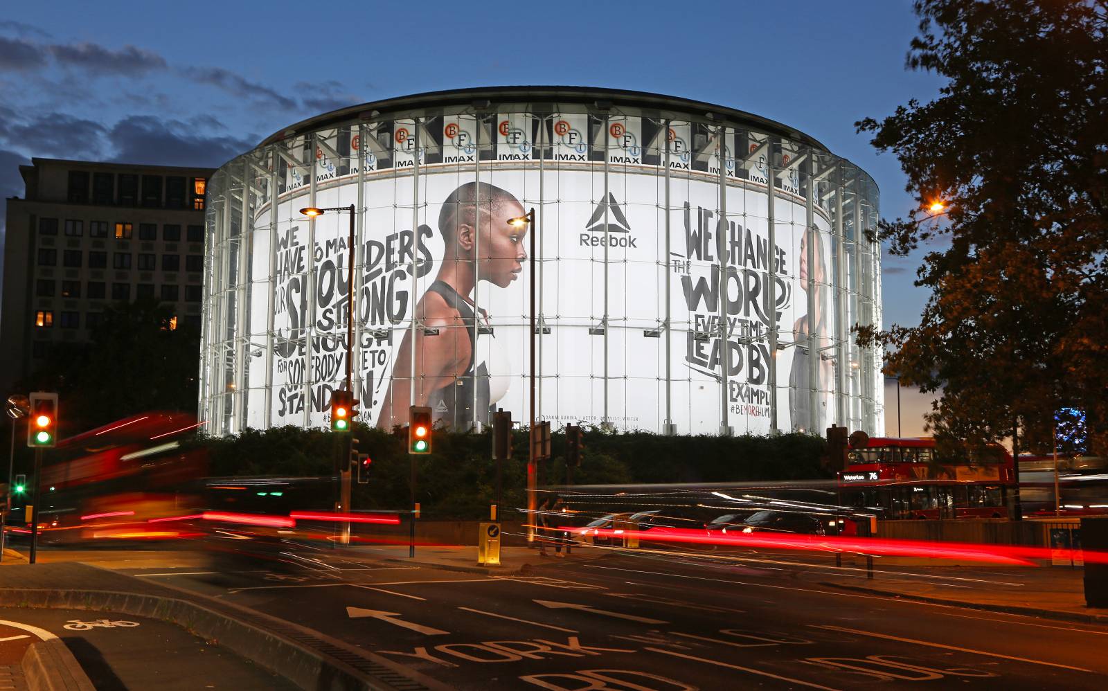 The IMAX with transparent walls displays an advertisement illuminated by LEDs, featuring an athletic woman in profile, promoting Reebok. Surrounding streets are busy with moving vehicles, and traffic lights are visible. The scene is at twilight.