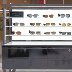 A display shelf in a store showcasing various Oliver Peoples sunglasses, neatly arranged in rows. The LED-lit sunglasses display highlights the different frame styles and lens tints perfectly. A sign indicating the brand is prominently placed at the top left.