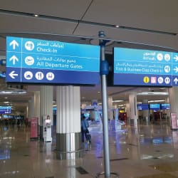 A spacious airport terminal with directional signs overhead. The blue signs display arrows and symbols indicating areas for check-in, departure gates, and fast-track check-in. Featuring English and Arabic text, DXB Airport chooses Bright Green Technology for new wayfinding signage in the bright, modern hall.