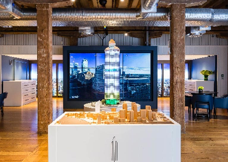 A detailed scale model of a modern skyscraper stands on a white pedestal in an industrial-themed room with exposed beams and ducts. Behind the model, a large screen displays an aerial cityscape illuminated by architectural LED lighting. Tables and chairs are arranged around the room, and potted plants are visible.