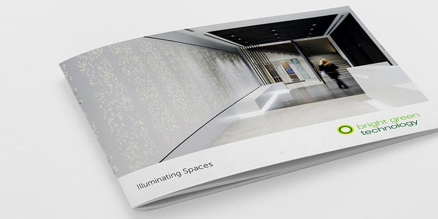 A white brochure titled "Illuminating Spaces" is partially open, revealing a modern, minimalistic interior with white walls, flooring, and sleek furniture. The logo and name "Bright Green Technology" are visible on the bottom right corner of the brochure's cover.