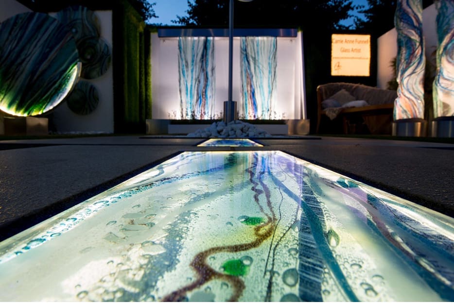 A glowing walkway with intricate, illuminated glass art leads to a display of vertical glass panels with flowing, blue-green patterns. The surrounding area features additional LED glass artworks, all set under a darkening evening sky.