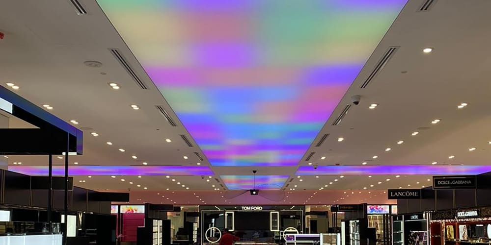 A high-end retail store ceiling adorned with colorful, gradient LED light sheet panels. The store features counters from luxury brands like Tom Ford, Lancome, and Dolce & Gabbana, creating a sophisticated shopping environment. Bright white spotlights are also evenly distributed throughout the space.