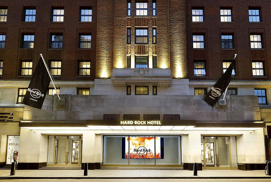 The exterior of Hard Rock Hotel at dusk, showcasing its illuminated signage and entrance. Black flags with the Hard Rock logo flank the entrance. The building's brick facade is accented by LED illumination, highlighting its architectural details.