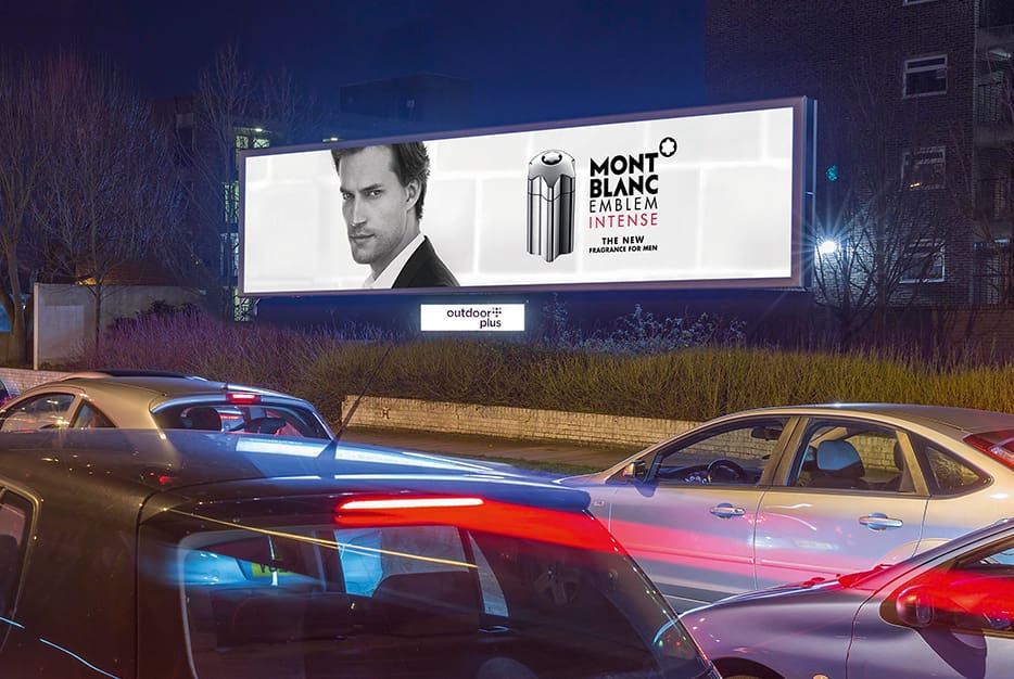 A brightly lit Mont Blanc billboard ad, powered by LEDs , is displayed on the side of the road at night, featuring an image of a man and advertising the 'Emblem Intense' fragrance. Several cars are stopped at a traffic light in front of the billboard.