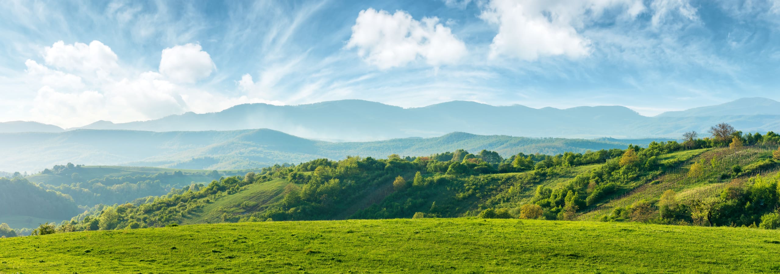 A panoramic view of a lush, green landscape with rolling hills and distant mountains under a bright blue sky dotted with scattered clouds. The foreground features a grassy field, while the midground has trees and shrubs.