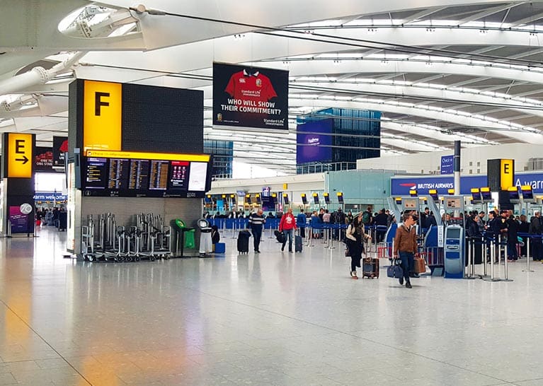 A spacious airport terminal with multiple check-in counters and a digital flight information board. Heathrow departures signs, LED illuminated by Bright Green Technology, cast a vibrant glow. People stand in lines, some using kiosks. Baggage carts and a large ceiling with arching beams are visible.