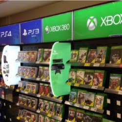 A store display showcases video games for various consoles, including PS4, PS3, Xbox 360, and Xbox One. Shelves are filled with game cases neatly organized, enhanced by custom-shaped LED light panels used for Tesco Gaming displays highlighting each console logo prominently above each shelf section.