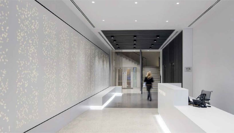 A spacious, modern office reception area with a sleek white desk to the right and a large LED light panel wall with a dotted pattern to the left. A blurred figure is walking towards a staircase in the back, leading to an upper floor. Minimalistic and well-lit setting.