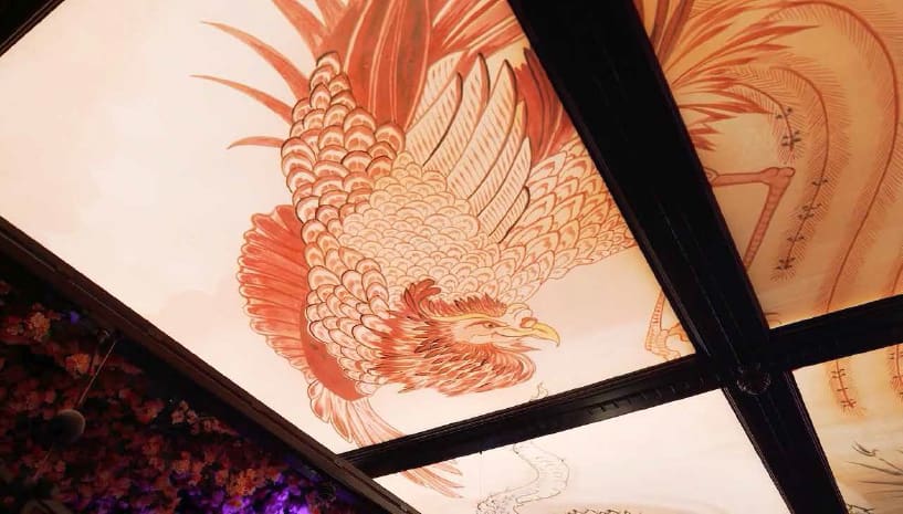 A ceiling decorated with a detailed illustration of a large bird, resembling a phoenix, in shades of brown and beige is subtly enhanced by an LED light panel. The artwork is divided into sections by dark wooden beams and surrounded by additional ornate decorations.