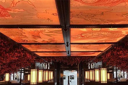 A richly decorated indoor restaurant with LED illuminated ceiling panels depicting intricate dragon designs. The walls are adorned with hanging red blossoms, and warm yellow lighting casts a cozy glow over the seating arrangement and tables.