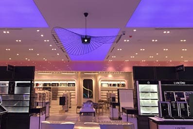 An elegant, modern retail store interior with a purple-lit ceiling. The store features neatly arranged shelves and display cases, all illuminated by spotlights. A large, contemporary light fixture and LED headlit shopfloor displays add to the vibrant ambiance.