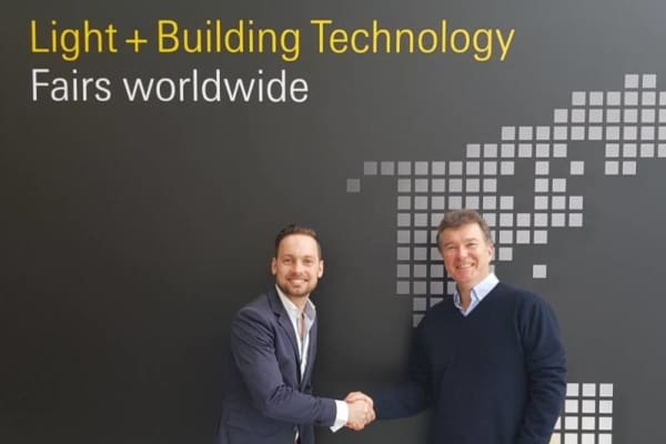 Two men in business attire shake hands in front of a wall displaying the text "Light + Building Technology Fairs worldwide" alongside a pixelated world map graphic.