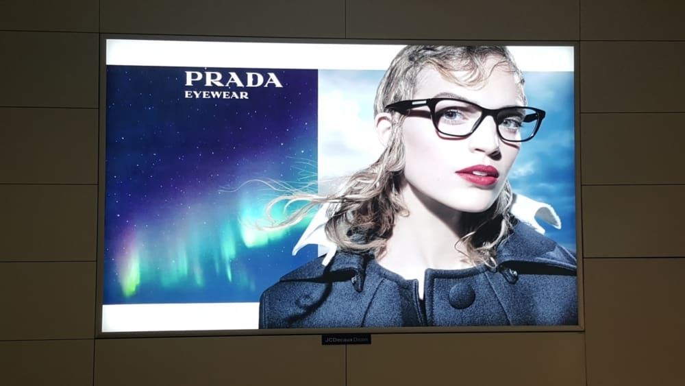 Advertisement for Prada eyewear featuring a person wearing black-framed glasses and a stylish coat. The background showcases a vibrant aurora borealis against a night sky, enhanced by LED backlit graphics. The Prada logo is at the top left with the word "EYEWEAR" beneath it—perfect for out of home displays.