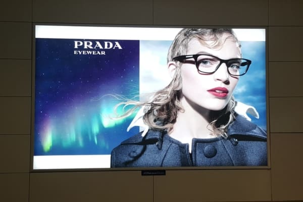 Advertisement for Prada eyewear featuring a person wearing black-framed glasses and a stylish coat. The background showcases a vibrant aurora borealis against a night sky, enhanced by LED backlit graphics. The Prada logo is at the top left with the word "EYEWEAR" beneath it—perfect for out of home displays.