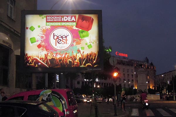 A large billboard lit up at night showcasing "Beer Fest" with vibrant LED backlit graphics and a crowd of people raising their hands. The surrounding area features buildings, a Canon sign, and parked cars, capturing the essence of out of home advertising as pedestrians cross the street.