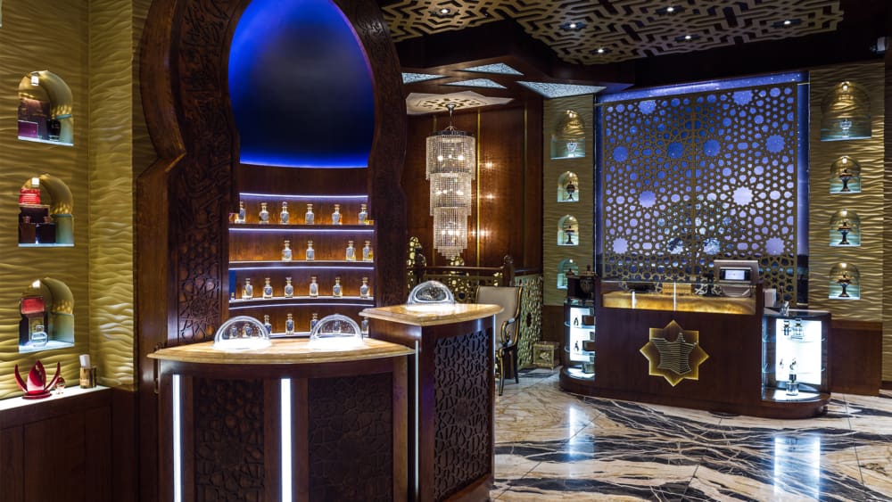 A luxurious perfume store features intricate woodwork, arched display niches with illuminated perfume bottles and LED shelf lighting, a central counter with glass domes, and a patterned backlit wall. The floor has a sleek, polished marble finish. Text credits TGP Interiors & Arabian Oud.