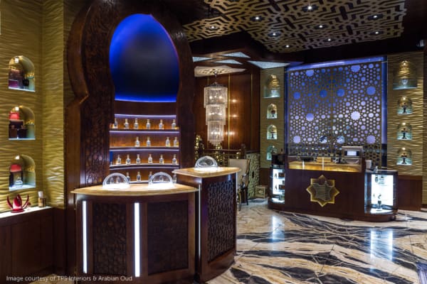 A luxurious perfume store features intricate woodwork, arched display niches with illuminated perfume bottles and LED shelf lighting, a central counter with glass domes, and a patterned backlit wall. The floor has a sleek, polished marble finish. Text credits TGP Interiors & Arabian Oud.