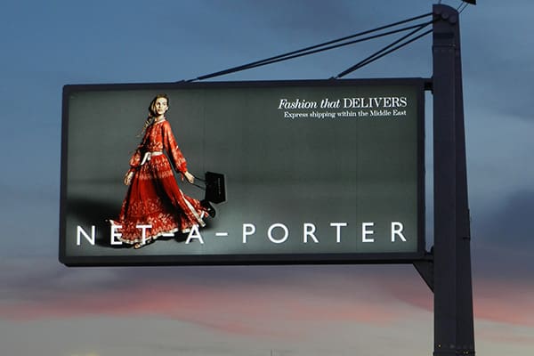 A billboard with LED backlit graphics displays an advertisement for NET-A-PORTER, featuring a woman in an elegant red dress carrying a black handbag. The text reads, "Fashion that DELIVERS. Express shipping within the Middle East." The sunset hue of the sky adds to this striking out-of-home display.