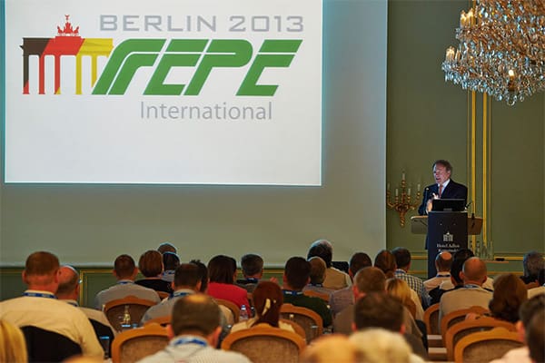 A conference room filled with attendees facing a podium where a speaker is presenting. Behind him is a large screen displaying the text "BERLIN 2013 FEPE International" with an accompanying logo. A chandelier hangs from the ceiling, adding elegance to the setting.