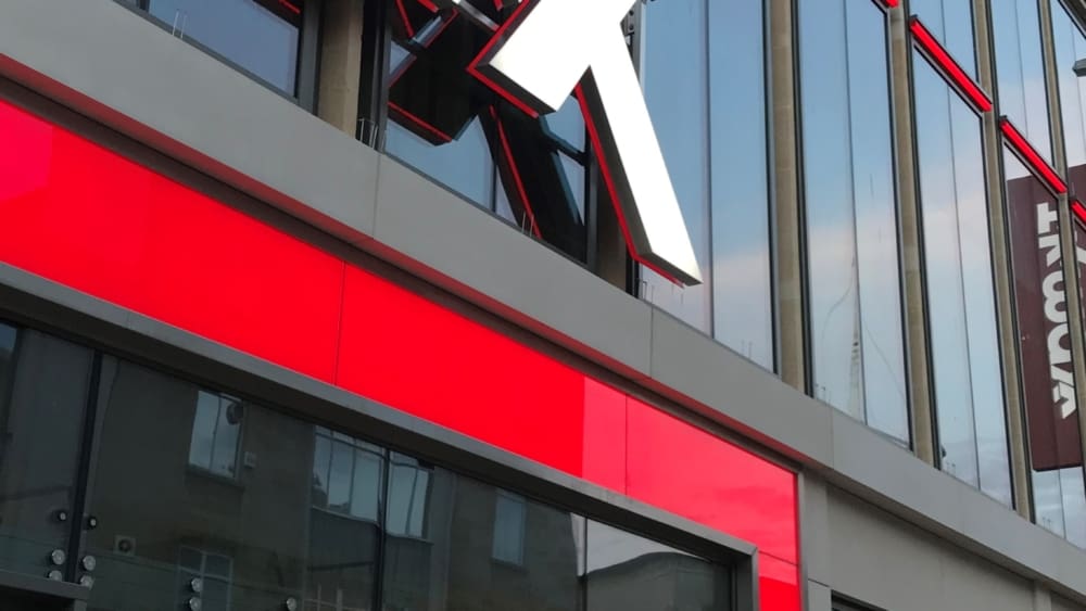 A large retail store with the red and white TJ Maxx logo on the exterior. The front features large glass windows revealing vibrant store displays and entrance doors. The building is modern with reflective windows, brand illumination through an LED light sheet panel, and a red accent around the entrance.