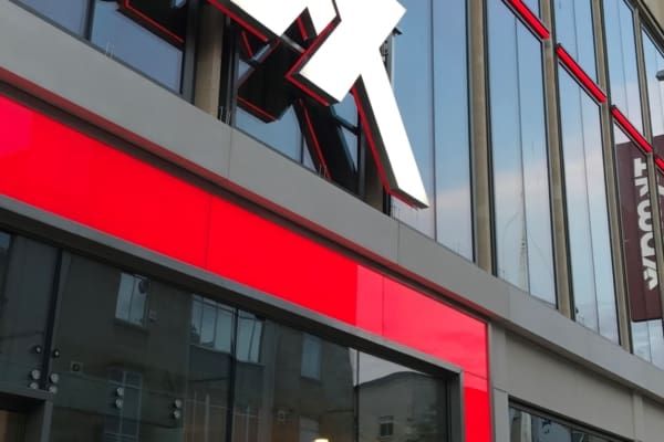 A large retail store with the red and white TJ Maxx logo on the exterior. The front features large glass windows revealing vibrant store displays and entrance doors. The building is modern with reflective windows, brand illumination through an LED light sheet panel, and a red accent around the entrance.