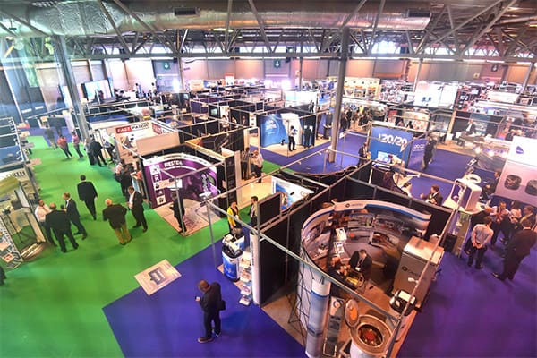 A bustling trade show floor with numerous booths displaying various products and services. Attendees are walking around, engaging with exhibitors, and exploring the different displays. The floor is covered in sections of green and blue carpet, and the venue is well-lit.