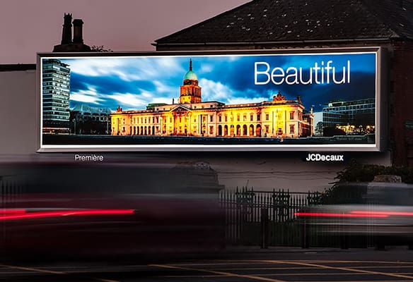 A large billboard at dusk displays a brightly lit image of a historic building with a domed roof and classical architecture under the word "Beautiful." Utilizing LED backlit graphics, the name "JCDecaux" is visible on the bottom right. Blurred red and white car lights are in the foreground.