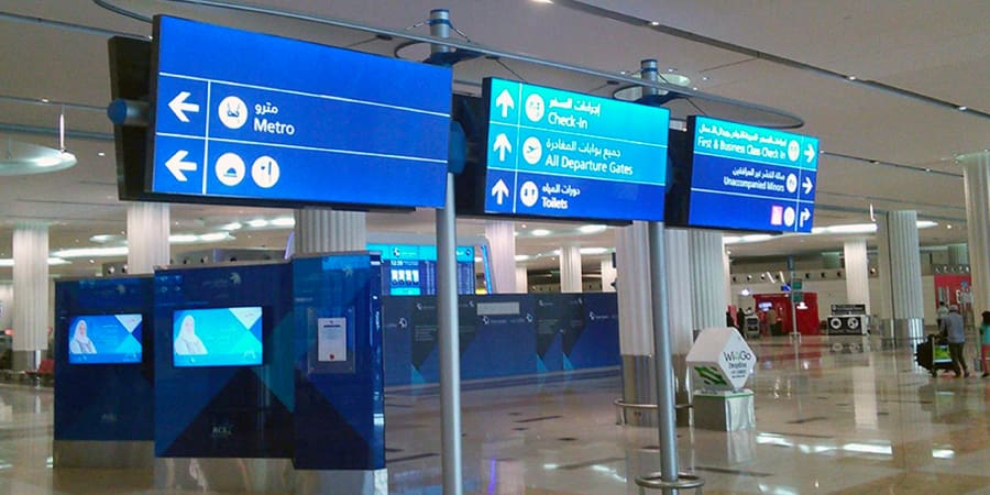 Digital signs with white text on a blue background in both Arabic and English show directions to the Metro, Check-in, All Departure Gates, Toilets, Arrivals, Baggage Reclaim, Transfers, and Meet and Assist. The area has pillars and a reflective floor illuminated by an advanced LED wayfinding signage system.