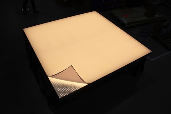 A large light panel is illuminated on a dark surface. One corner is peeled back, revealing a grid of small LED lights underneath. The surrounding area is dimly lit.